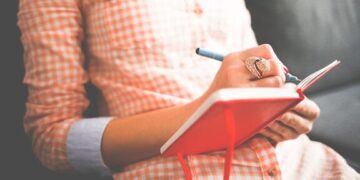 Person in a plaid shirt writing in a red notebook planning tasks, Master Your Website Maintenance Schedule: Boost Performance Now!